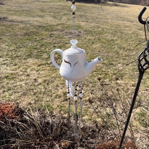 Puede incluir: Un carillón de viento de tetera de cerámica blanca con un diseño decorativo. La tetera está suspendida por una cadena con cuentas y plumas colgando debajo. El fondo presenta un campo cubierto de hierba y un edificio pequeño.