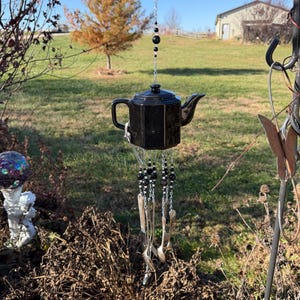 Puede incluir: Un carillón de viento de tetera de cerámica negra colgando al aire libre. La tetera está suspendida por una cadena con cuentas negras. Los utensilios de plata cuelgan debajo de la tetera, creando un adorno de jardín único y decorativo.