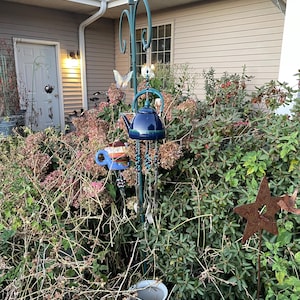 Puede incluir: Un carillón de tetera azul con cuentas y una mariposa colgando de un soporte de metal en un jardín.
