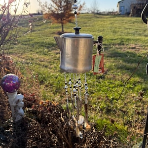 Puede incluir: Un carillón hecho a mano con una cafetera de aluminio vintage, cucharas y cuentas decorativas. El carillón cuelga de una cadena y tiene una etiqueta roja. El fondo incluye un campo de hierba y árboles.