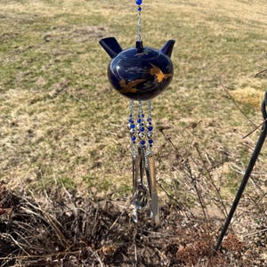 Puede incluir: Un carillón de viento con forma de tetera azul marino con detalles dorados de pájaros. La tetera cuelga de una cadena con cuentas azules y doradas, y cubiertos cuelgan debajo. El carillón de viento está suspendido al aire libre.