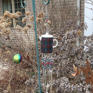 Puede incluir: Una tetera blanca con un patrón de cuadros cuelga de un soporte de metal con una cadena de cucharas de plata y cuentas rojas y azules. La tetera es un carillón de viento.