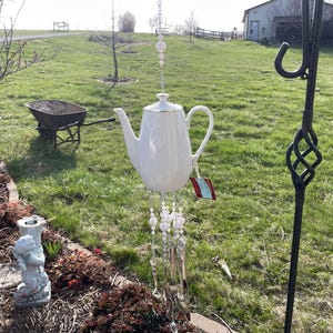 Puede incluir: Carillón de viento de porcelana blanca en forma de tetera con una cadena plateada y cuentas de perlas. El carillón está colgado de un gancho de metal negro.