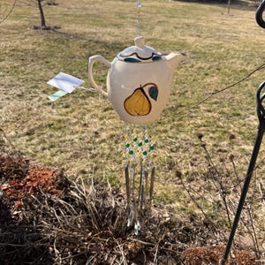 Puede incluir: Carrillón de viento con forma de tetera crema con diseño de pera. La tetera tiene un asa y un pico, suspendida por una cadena con cuentas de colores. El carillón de viento tiene varillas de vidrio y está colgado al aire libre.