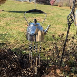 Puede incluir: Un carillón hecho a mano con una tetera plateada brillante. La tetera cuelga de una cadena con cuentas negras y transparentes. Cucharas y tenedores pequeños cuelgan de la tetera. El fondo muestra un jardín con césped y un edificio.