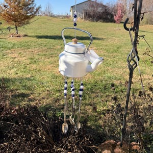 Puede incluir: Un carillón de viento con forma de tetera de cerámica blanca con ribete azul y un pomo de madera. El carillón cuelga de un gancho negro y presenta cuentas azules y blancas, con cucharas y tenedores colgando debajo. El fondo muestra un campo de hierba.
