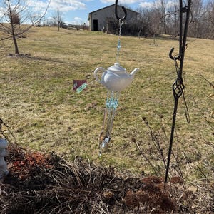 Puede incluir: Un carillón de viento de tetera de cerámica blanca colgando de un gancho de pastor de metal negro. El carillón de viento tiene cuentas azules y campanillas de metal. El fondo muestra un campo cubierto de hierba, un edificio y un cielo parcialmente nublado.