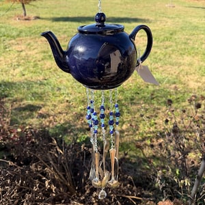 Puede incluir: Un carillón de viento con forma de tetera de cerámica azul marino. La tetera cuelga de una cadena de cuentas azules y plateadas. Debajo cuelgan cubiertos, incluyendo tenedores y cucharas, creando un adorno de jardín único.