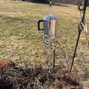 Puede incluir: Un carillón hecho a mano con una cafetera de aluminio vintage. La cafetera tiene un asa negra y está suspendida por una cadena con cuentas negras y plateadas. Varillas de vidrio transparente y cuentas negras cuelgan debajo.