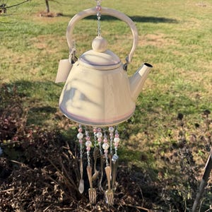 Puede incluir: Carrillón de viento con forma de tetera crema con asa y boquilla. La tetera está decorada con rayas rosas y grises. Debajo de la tetera cuelgan cubiertos de plata y cuentas rosas y blancas. El carillón está suspendido de una cadena con cuentas rosas y transparentes.