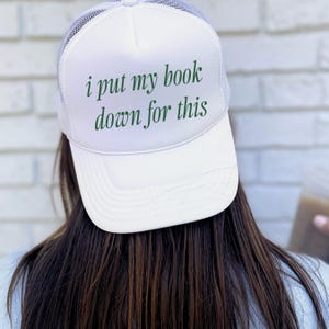 May include: A white trucker hat with a mesh back and a solid front panel. The front features the dark green text "i put my book down for this" printed in a serif font. The hat has a structured crown and a curved brim, providing a classic look.