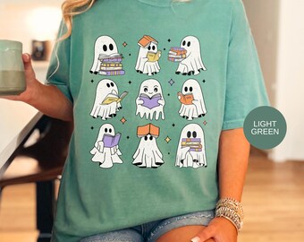 May include: A light green t-shirt featuring a graphic print of nine white ghosts, each interacting with colorful books. The ghosts are depicted in various poses, some reading, some holding stacks of books, and others with books balanced on their heads.