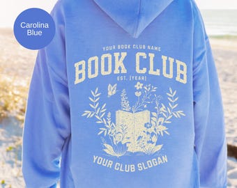 May include: A Carolina blue hooded sweatshirt featuring a cream-colored graphic on the back. The design includes an open book surrounded by botanical illustrations, with the text "YOUR BOOK CLUB NAME", "BOOK CLUB", "EST. (YEAR)", and "YOUR CLUB SLOGAN".