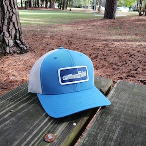 May include: A blue baseball cap with a white mesh back. The front of the cap features a rectangular patch with the text "CHESAPEAKE BAY" and a boat graphic. The cap is sitting on a wooden surface.