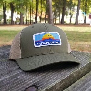 May include: A green and tan trucker hat with a white patch that says "CHESAPEAKE BAY" and features a sunset, a boat, and birds.