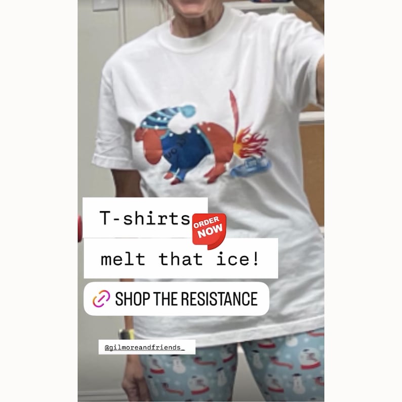 May include: White t-shirt featuring a colorful cartoon illustration of a creature melting ice. The text "melt that ice!" is displayed below the image. The shirt also has the text "SHOP THE RESISTANCE".