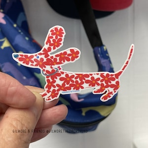 May include: A white dachshund sticker with a red floral pattern. The sticker says "Gilmore & Friends" on the bottom.