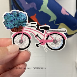 May include: Pink bicycle with a basket full of blue flowers. The bicycle has black wheels and a black seat. The words "Gilmore & Friends" are written on the back tire.