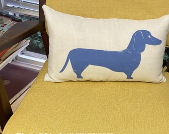 May include: A rectangular cream-colored pillow with a blue dachshund illustration. The pillow is placed on a yellow chair. The text "Gilmore & Friends @GILMOREANDFRIENDS_" is at the bottom of the image.