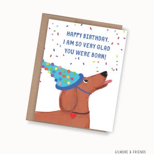 May include: A birthday card featuring a brown dachshund wearing a blue party hat with colorful polka dots. The card reads "HAPPY BIRTHDAY. I AM SO VERY GLAD YOU WERE BORN!" with confetti. The card is inside a brown envelope.