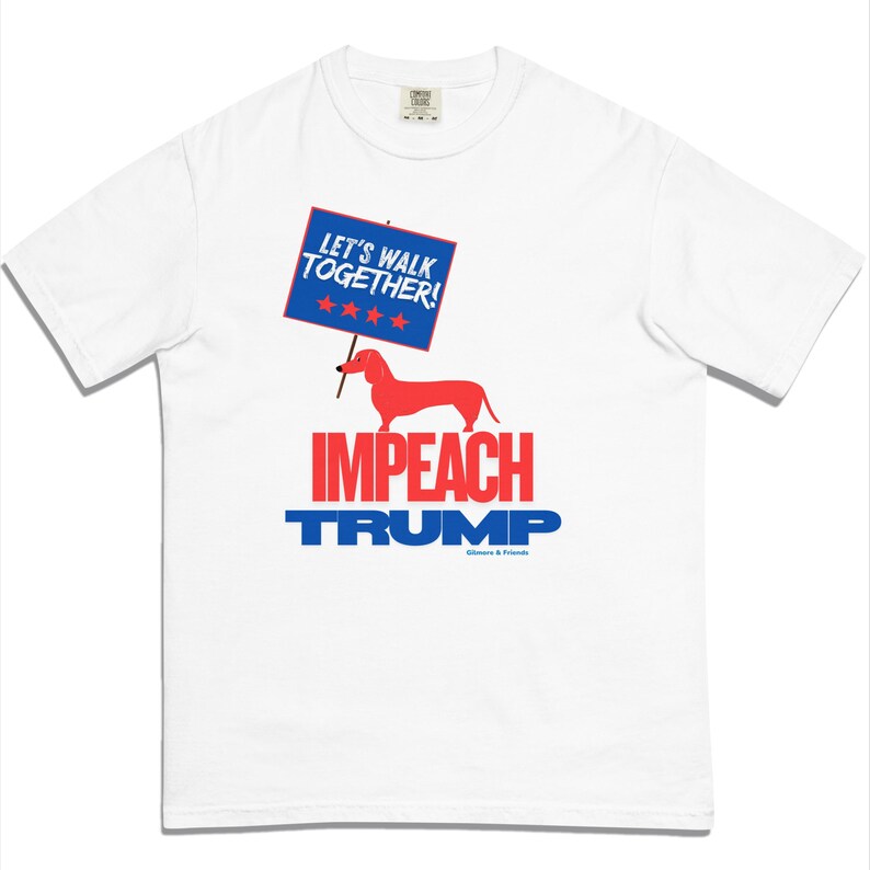 May include: White t-shirt with a graphic design. The design features a red dachshund holding a sign that reads "LET'S WALK TOGETHER!" above the words "IMPEACH TRUMP" in red and blue. The shirt is from Gilmore & Friends.