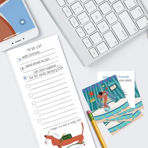 May include: A white to-do list with a check mark next to "walk Gilmore", "send email to list", and "call card supplier for NY cards delivery ETA". The list is on a white surface with a keyboard, a yellow pencil, and a stack of business cards with a cartoon dog illustration.