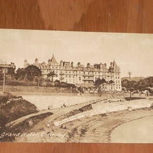 May include: A black and white vintage postcard featuring a photo of the Grand Hotel in Conway, Wales. The hotel is a large, multi-story building with a prominent tower. The postcard is slightly faded and has a vintage look.