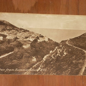May include: A black and white vintage postcard showing a view of the town of Lynton from Summerhouse Hill. The postcard features a winding road leading up the hill with a view of the town and the sea in the distance.