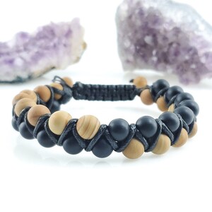 Puede incluir: Una pulsera de doble hebra con cuentas de madera negras y marrón claro alternadas, unidas por un cordón negro. La pulsera se exhibe sobre una superficie blanca, con un cristal púrpura en el fondo.