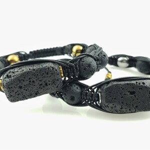 May include: Two black lava stone bracelets with gold beads and black cord. The bracelets are knotted and feature a rectangular lava stone bead.