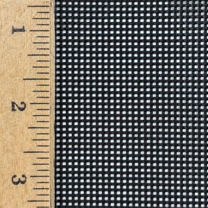 May include: A black mesh fabric with a uniform grid pattern positioned next to a wooden ruler. The ruler shows a scale in inches, with the mesh covering the area from the 1-inch mark to the 3-inch mark, displaying the fine square weave.
