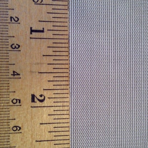 May include: A light grey, finely woven fabric with a consistent, tight mesh texture. A wooden ruler is positioned vertically along the left edge, showing markings in inches, with the fabric extending past the three-inch mark on the ruler.