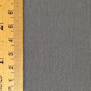 May include: A piece of grey fabric with a fine, uniform knit texture positioned next to a wooden ruler. The ruler shows the fabric width spans approximately 6.4 cm, displaying clear vertical lines and a consistent, dense weave pattern throughout.