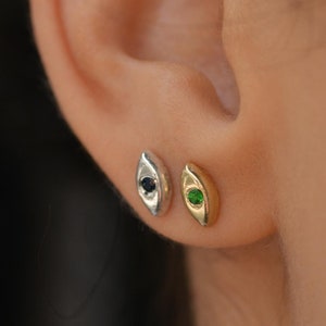 Może przedstawiać: Trzy złote, srebrne i białe kolczyki w kształcie oka z małymi kamieniami szlachetnymi. Kolczyki mają kształt oka z małym kamieniem szlachetnym w środku. Kamienie szlachetne są czerwone, zielone i czarne.