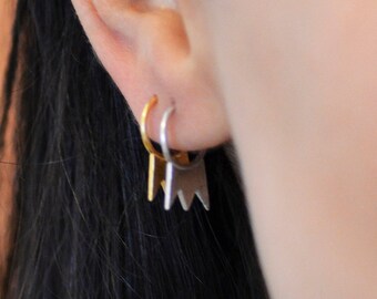 May include: Close-up of an ear with three earrings. The top earring is a small silver hoop. The middle earring is a small gold hoop. The bottom earring is a silver geometric design.