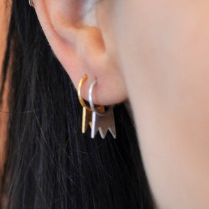 May include: Close-up of an ear with three earrings. The top earring is a small silver hoop. The middle earring is a small gold hoop. The bottom earring is a silver geometric design.