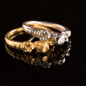 Pode incluir: Dois anéis, um dourado e outro prateado, empilhados sobre uma superfície preta reflexiva. O anel dourado tem uma banda texturizada e uma pedra central facetada. O anel prateado tem um design semelhante. Ambos os anéis estão focados.