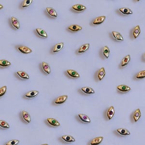 Może przedstawiać: Asortyment małych zawieszek w kształcie oka w odcieniach złota i srebra, każda z kolorowym kamieniem szlachetnym pośrodku. Kamienie szlachetne są w różnych kolorach, w tym różowym, zielonym, czerwonym i niebieskim. Zawieszki są rozrzucone na białej powierzchni.