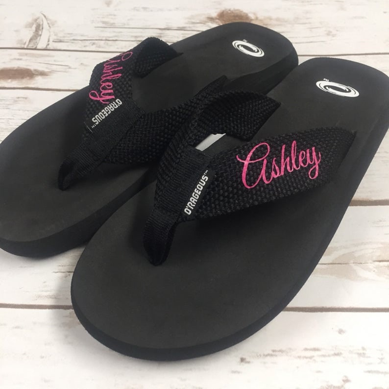 May include: A pair of black flip-flops with a black and white strap. The strap is embroidered with the name "Ashley" in pink.