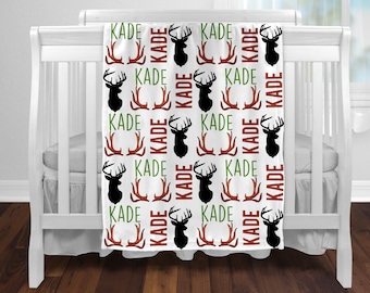 May include: White baby blanket with a repeating pattern of brown antlers, black deer heads, and the name "KADE" in green and red.