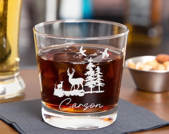 Puede incluir: Un vaso de rocas de vidrio transparente con un ciervo, dos árboles y dos pájaros grabados en el lateral. El vaso está lleno de líquido marrón y cubitos de hielo. El nombre "Carson" está grabado debajo del diseño.