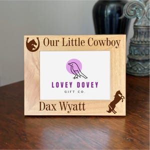 Op de afbeelding: Een lichtgekleurde houten fotolijst met de tekst "Our Little Cowboy" en "Dax Wyatt". De lijst heeft een vogel-logo en een silhouet van een steigerend paard. De lijst staat op een donker houten oppervlak.