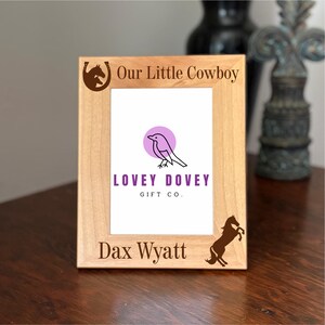 Op de afbeelding: Een lichtgekleurde houten fotolijst met de tekst "Our Little Cowboy" en "Dax Wyatt". De lijst heeft een logo met een vogel en de woorden "LOVEY DOVEY GIFT CO.", samen met een paard en hoefijzer.