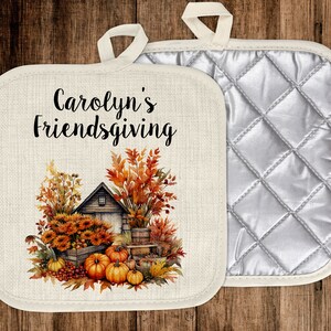 Puede incluir: Un agarrador de olla de algod&oacute;n blanco con un respaldo acolchado. La parte delantera presenta una ilustraci&oacute;n de oto&ntilde;o de una casa, calabazas y girasoles. El texto "Carolyn's Friendsgiving" est&aacute; impreso en negro.