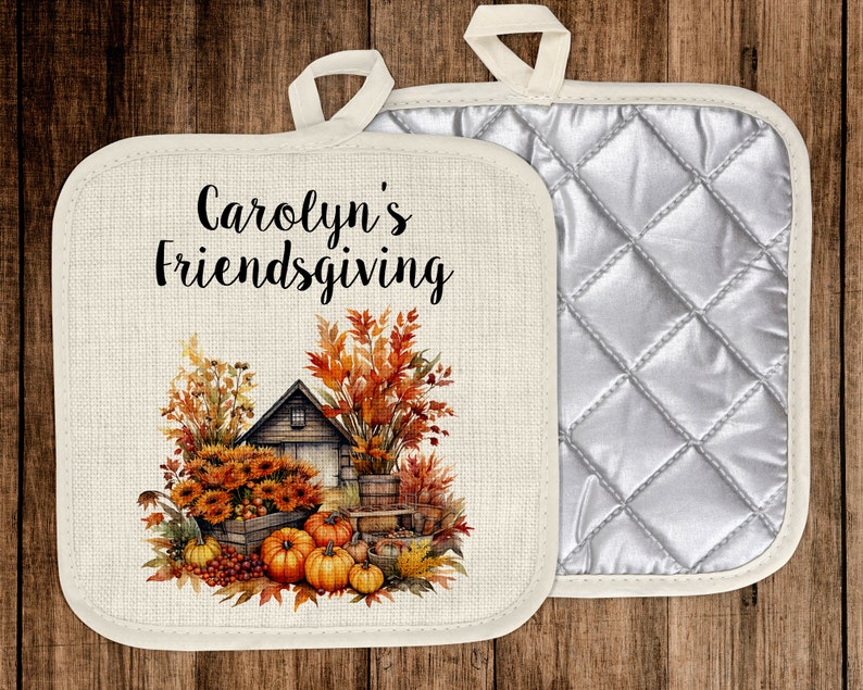 Puede incluir: Un agarrador de olla de algod&oacute;n blanco con un respaldo acolchado. La parte delantera presenta una ilustraci&oacute;n de oto&ntilde;o de una casa, calabazas y girasoles. El texto "Carolyn's Friendsgiving" est&aacute; impreso en negro.