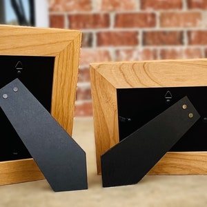 Op de afbeelding: Twee houten fotolijsten met een lichtbruine afwerking. De lijsten hebben een zwarte achterkant en een zwarte ezelstandaard. De lijsten worden gepresenteerd tegen een bakstenen muur.
