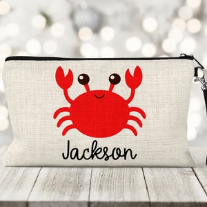 May include: A white canvas zippered pouch with a red cartoon crab and the name "Jackson" printed on the front. The pouch has a black zipper and a black strap.