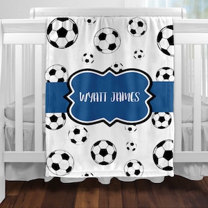 May include: White baby blanket with a blue ribbon and a blue frame with the name "WYATT JAMES" printed on it. The blanket is decorated with a pattern of black and white soccer balls.
