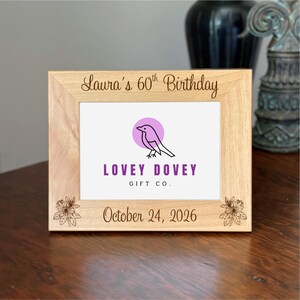 May include: A light-colored wooden picture frame with the text "Laura's 60th Birthday" and "October 24, 2026". The frame features a logo with a bird inside a purple circle and the words "LOVEY DOVEY GIFT CO."