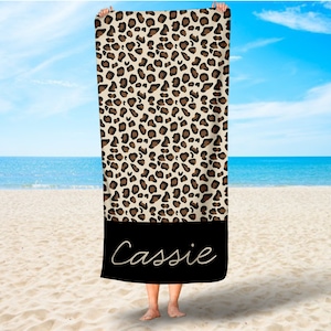 May include: A beach towel with a leopard print design and a black border. The name "Cassie" is written in white script on the black border.